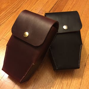 May include: Two leather cases, one burgundy and one black, with rounded flap closures secured by metal snaps. The cases are rectangular with white stitching along the edges. The cases are likely designed to hold small items.