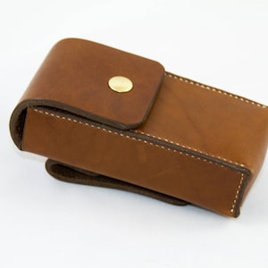 May include: A brown leather case with a brass button closure. The case is designed to hold a small item, such as a pen or a knife.