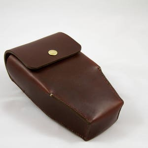 May include: A rectangular, dark brown leather case with a rounded top flap secured by a gold-colored snap. The case has a smooth, polished finish and appears to be handmade. The background is plain white.