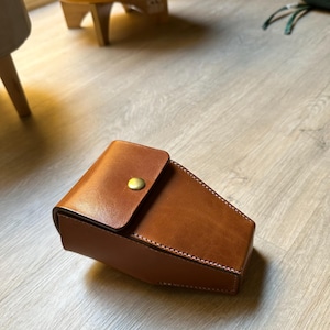 May include: A brown leather case with a flap closure secured by a gold-colored button. The case has visible stitching around the edges and a smooth, polished surface. The case is designed to hold a small item.