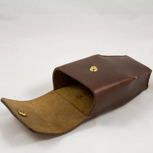 May include: A brown leather case with a flap that opens to reveal a lighter brown interior. The case has a gold-colored snap closure on the front and a matching snap on the flap. The case is likely for storing small items.