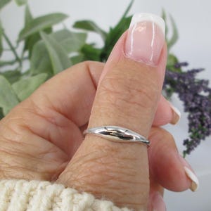 Op de afbeelding: Een zilveren ring met een glad, langwerpig ontwerp wordt aan een vinger gedragen. Het gepolijste oppervlak van de ring reflecteert licht. De achtergrond omvat groen en een witte trui.