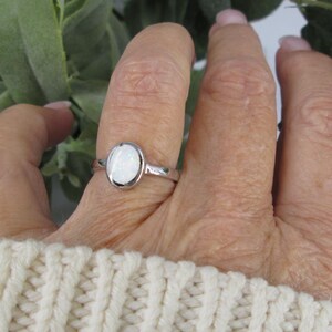 May include: A silver ring with an oval, white gemstone is displayed on a hand. The ring's band is thin and polished. The gemstone is set in a bezel setting. The hand is wearing a cream-colored sweater.