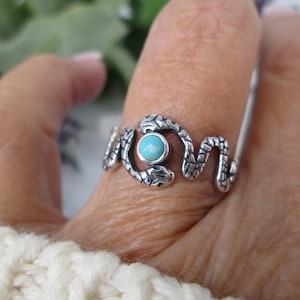May include: A silver ring featuring a snake design with a turquoise stone. The ring wraps around the finger with the snake's head and tail meeting on either side of the turquoise stone. The ring is worn on a finger.