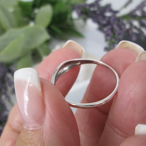 Op de afbeelding: Een gepolijste zilveren ring met een gladde band. De ring wordt tussen vingers met verzorgde nagels gehouden. De achtergrond bevat groen gebladerte en paarse bloemen. Het ontwerp van de ring is eenvoudig en elegant.