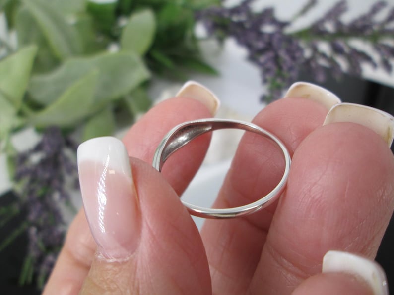 Op de afbeelding: Een gepolijste zilveren ring met een gladde band. De ring wordt tussen vingers met verzorgde nagels gehouden. De achtergrond bevat groen gebladerte en paarse bloemen. Het ontwerp van de ring is eenvoudig en elegant.