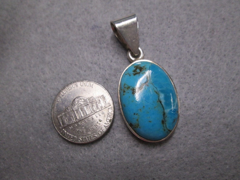 Puede incluir: Un colgante de plata con una piedra turquesa azul. La piedra es de forma ovalada y tiene un color azul claro con vetas azules m&aacute;s oscuras. El colgante es aproximadamente del tama&ntilde;o de una moneda de un c&eacute;ntimo de euro.