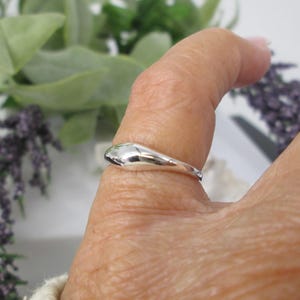 Op de afbeelding: Een gepolijste zilveren ring met een glad, druppelvormig ontwerp. De ring wordt getoond op een vinger, met een vage achtergrond van groen en paarse bloemen. Het oppervlak van de ring reflecteert licht.