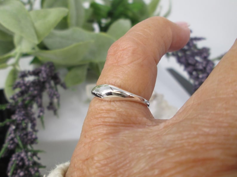 Op de afbeelding: Een gepolijste zilveren ring met een glad, druppelvormig ontwerp. De ring wordt getoond op een vinger, met een vage achtergrond van groen en paarse bloemen. Het oppervlak van de ring reflecteert licht.