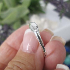 Op de afbeelding: Een zilveren ring met een druppelvormig ontwerp. De ring wordt vastgehouden in een hand met verzorgde nagels. Het oppervlak van de ring reflecteert licht, waardoor een heldere, glanzende uitstraling ontstaat. De achtergrond is wazig, met hints van groen en wit.