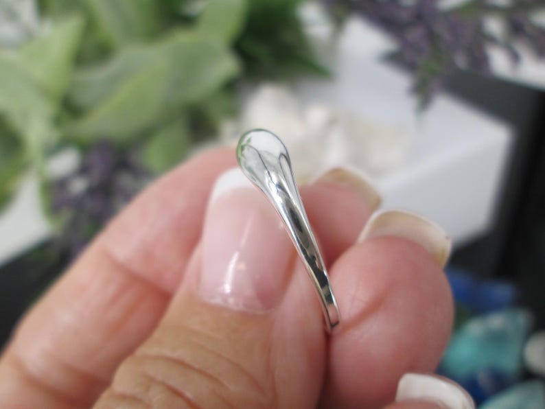 Op de afbeelding: Een zilveren ring met een druppelvormig ontwerp. De ring wordt vastgehouden in een hand met verzorgde nagels. Het oppervlak van de ring reflecteert licht, waardoor een heldere, glanzende uitstraling ontstaat. De achtergrond is wazig, met hints van groen en wit.