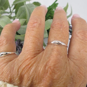 Op de afbeelding: Close-up van een hand met twee zilveren ringen. De ringen hebben een glad, gebogen ontwerp. De achtergrond bevat groen en paars gebladerte. De ringen zijn een eenvoudig, elegant accessoire.