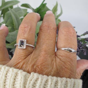 Op de afbeelding: Een hand met twee ringen. E&eacute;n ring heeft een rechthoekige, donkergekleurde steen omgeven door een halo van kleinere stenen. De andere ring is een eenvoudige, gebogen band. De hand draagt een cr&egrave;mekleurige trui.