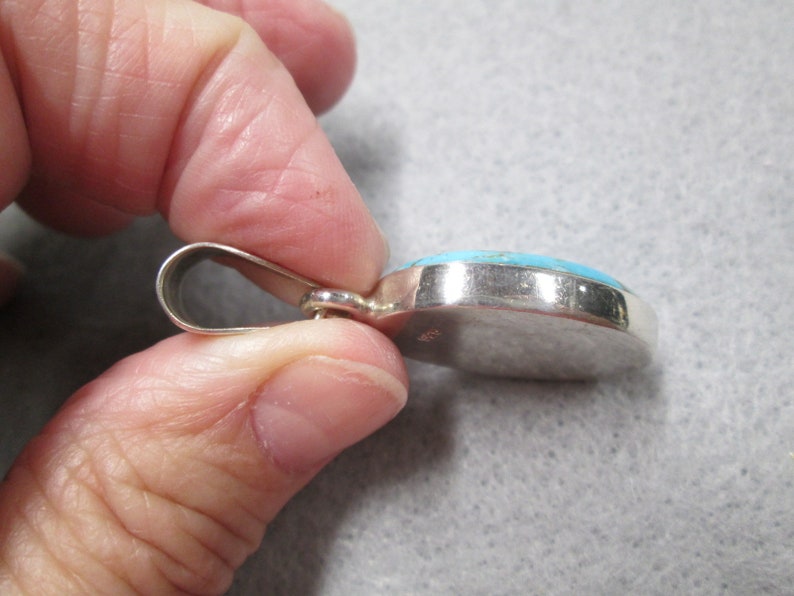 Puede incluir: Un colgante de plata con una piedra turquesa engastada en un bisel de plata. El colgante tiene un bucle para sujetarlo a una cadena.