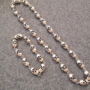 May include: A silver necklace and bracelet set with oval-shaped beads. The beads have a decorative, openwork design.