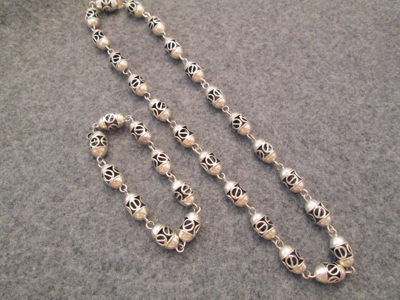 May include: A silver necklace and bracelet set with oval-shaped beads. The beads have a decorative, openwork design.