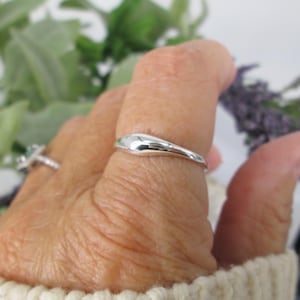 Op de afbeelding: Een glanzende, zilveren ring met een glad, druppelvormig ontwerp. De ring wordt aan een vinger gedragen, met een tweede ring zichtbaar op de achtergrond. De ring staat tegen een achtergrond van groen en paars gebladerte.