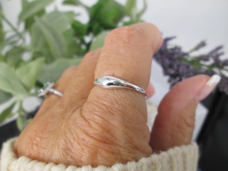 Op de afbeelding: Een glanzende, zilveren ring met een glad, druppelvormig ontwerp. De ring wordt aan een vinger gedragen, met een tweede ring zichtbaar op de achtergrond. De ring staat tegen een achtergrond van groen en paars gebladerte.