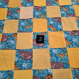 Peut inclure: Une courtepointe en patchwork compos&eacute;e de carr&eacute;s altern&eacute;s de tissu jaune &agrave; pois blancs et de tissu bleu sarcelle imprim&eacute; de lions et d'hippopotames de dessin anim&eacute; dans une jungle. Une petite &eacute;tiquette noire avec le chiffre 2 blanc est centr&eacute;e.