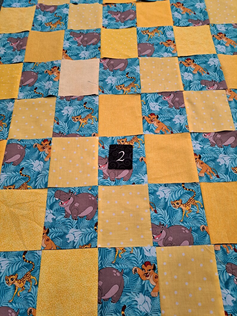 Peut inclure: Une courtepointe en patchwork compos&eacute;e de carr&eacute;s altern&eacute;s de tissu jaune &agrave; pois blancs et de tissu bleu sarcelle imprim&eacute; de lions et d'hippopotames de dessin anim&eacute; dans une jungle. Une petite &eacute;tiquette noire avec le chiffre 2 blanc est centr&eacute;e.
