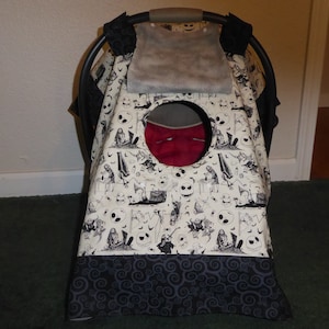 May include: A baby car seat cover with a cream-colored background featuring a black and white Halloween-themed pattern. The pattern includes skulls, bats, and other spooky imagery. The bottom border is a dark gray with a swirling design.