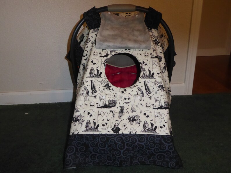 May include: A baby car seat cover with a cream-colored background featuring a black and white Halloween-themed pattern. The pattern includes skulls, bats, and other spooky imagery. The bottom border is a dark gray with a swirling design.