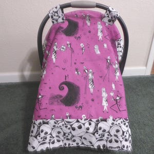 May include: A pink baby car seat cover featuring a black and white pattern of characters and imagery from The Nightmare Before Christmas. The bottom border is white with skull graphics. The car seat handle covers have a skull pattern.