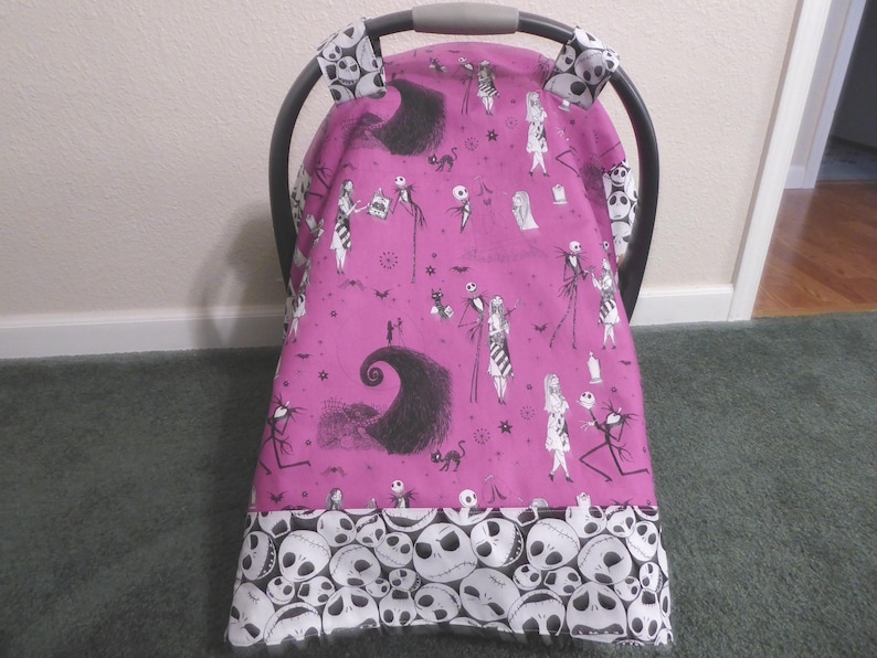 May include: A pink baby car seat cover featuring a black and white pattern of characters and imagery from The Nightmare Before Christmas. The bottom border is white with skull graphics. The car seat handle covers have a skull pattern.
