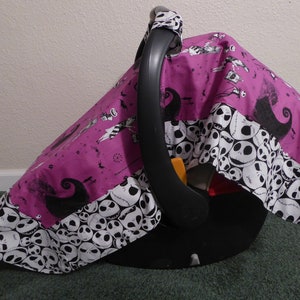 May include: A baby car seat with a custom cover featuring a purple background with black and white Jack Skellington characters and a border of Jack Skellington faces. The car seat has a black handle and base.