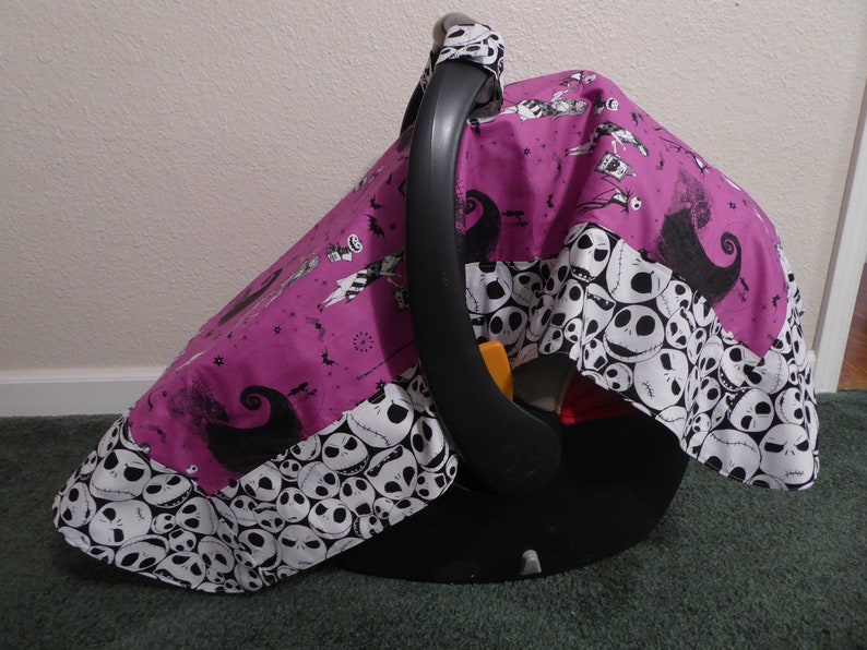 May include: A baby car seat with a custom cover featuring a purple background with black and white Jack Skellington characters and a border of Jack Skellington faces. The car seat has a black handle and base.