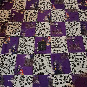 Peut inclure: Une courtepointe en patchwork compos&eacute;e de carr&eacute;s de tissu altern&eacute;s. Un motif pr&eacute;sente des visages blancs de Jack Skellington sur fond noir, tandis que l'autre montre Jack Skellington et Sally dans un cimeti&egrave;re violet. Une petite &eacute;tiquette noire avec le chiffre 1 est fix&eacute;e.