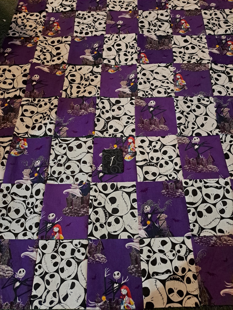 Peut inclure: Une courtepointe en patchwork compos&eacute;e de carr&eacute;s de tissu altern&eacute;s. Un motif pr&eacute;sente des visages blancs de Jack Skellington sur fond noir, tandis que l'autre montre Jack Skellington et Sally dans un cimeti&egrave;re violet. Une petite &eacute;tiquette noire avec le chiffre 1 est fix&eacute;e.