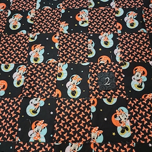 Puede incluir: Una colcha de retazos con cuadrados alternos de tela negra con un patr&oacute;n repetido de lazos naranjas y tela negra con un estampado de Minnie Mouse sobre un c&iacute;rculo azul claro y una luna naranja, con un peque&ntilde;o cuadrado negro etiquetado con un 2.