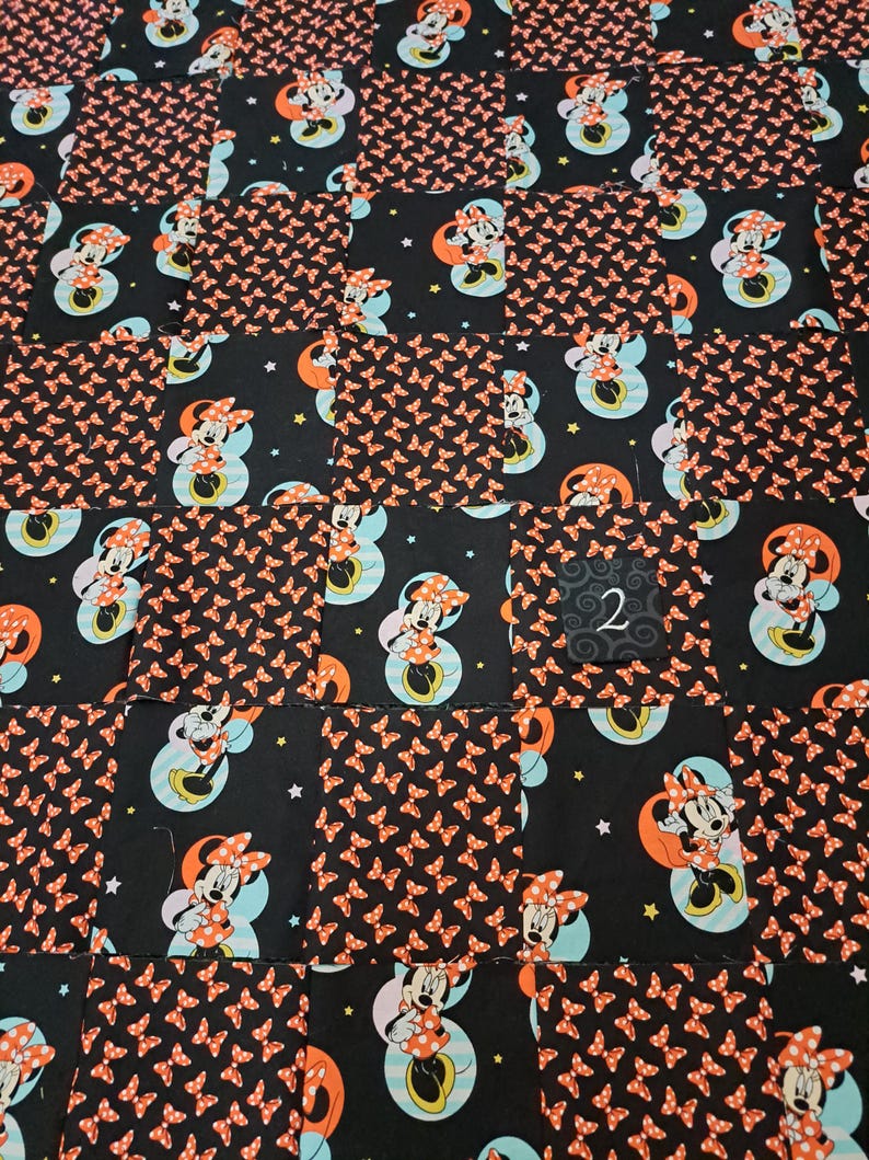Puede incluir: Una colcha de retazos con cuadrados alternos de tela negra con un patr&oacute;n repetido de lazos naranjas y tela negra con un estampado de Minnie Mouse sobre un c&iacute;rculo azul claro y una luna naranja, con un peque&ntilde;o cuadrado negro etiquetado con un 2.