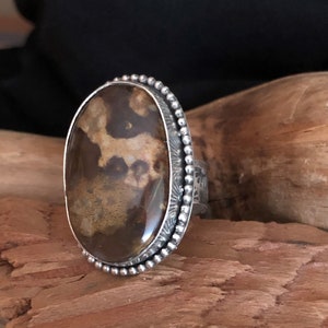 May include: Oval-shaped ring with a large, polished brown stone featuring lighter brown patterns. The stone is set in a silver band with a beaded edge and decorative details. The ring is displayed on a wooden surface.