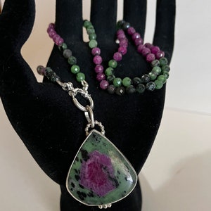 May include: A green and pink gemstone pendant necklace with a silver chain. The pendant is a teardrop shape and the necklace is made of small beads.