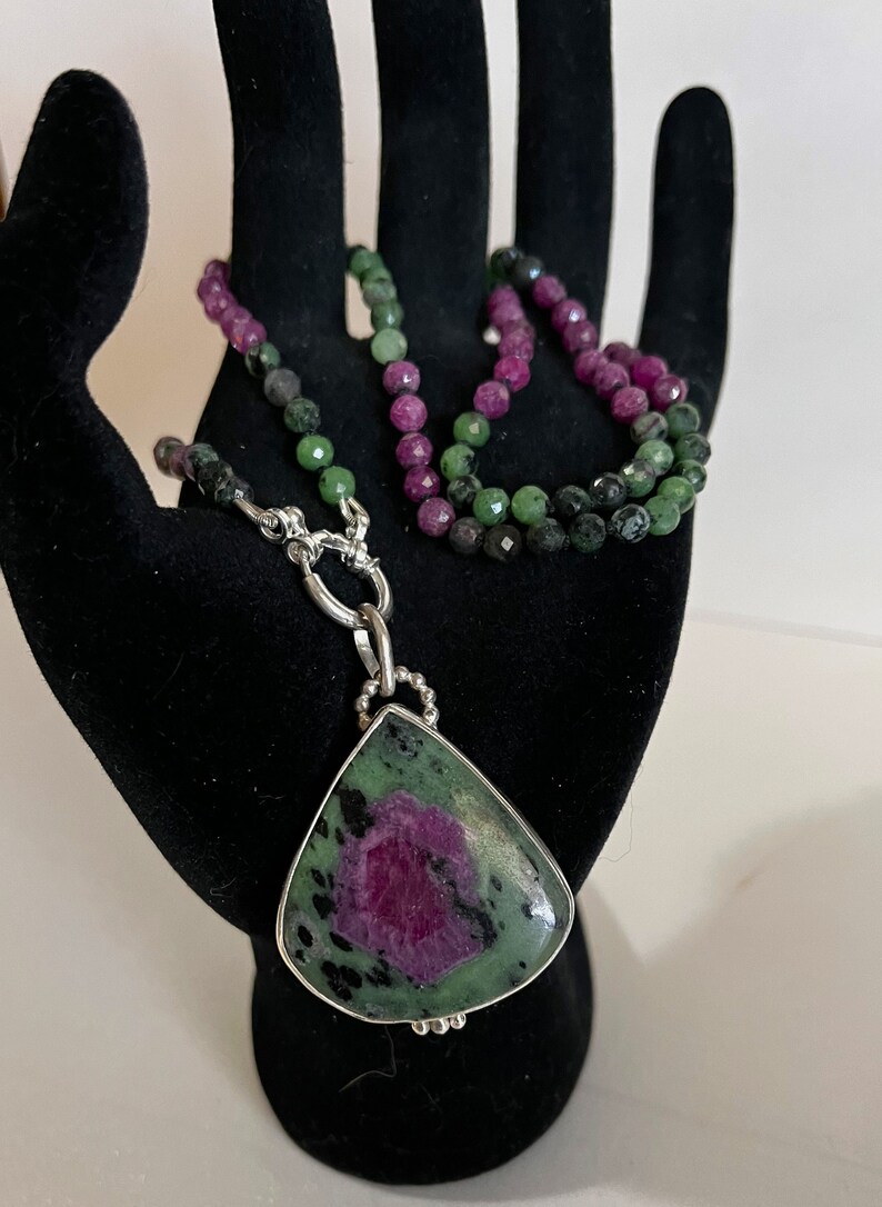 May include: A green and pink gemstone pendant necklace with a silver chain. The pendant is a teardrop shape and the necklace is made of small beads.