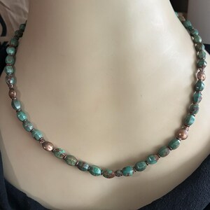 May include: A beaded necklace featuring oval-shaped turquoise-colored beads with brown marbling, interspersed with small, round, metallic copper-toned beads and two distinct copper-toned beads shaped like small fish with etched details.