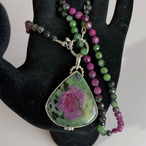 May include: A green and pink gemstone pendant necklace with a silver clasp. The pendant is a teardrop shape and has a green and pink color pattern. The necklace is made of small green and pink beads.
