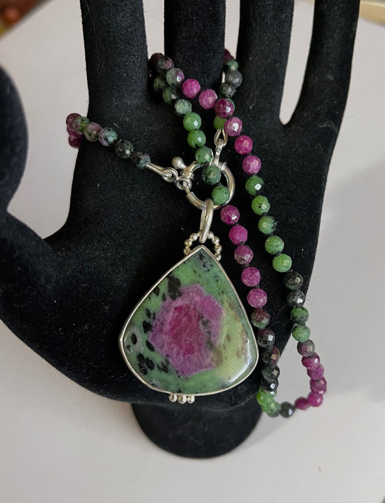 May include: A green and pink gemstone pendant necklace with a silver clasp. The pendant is a teardrop shape and has a green and pink color pattern. The necklace is made of small green and pink beads.
