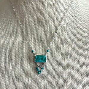 May include: A silver necklace featuring a rectangular teal stone pendant set in a serrated bezel. The pendant is accented with small dangling teal beads and is suspended from a delicate silver chain with elongated links and small teal beads.