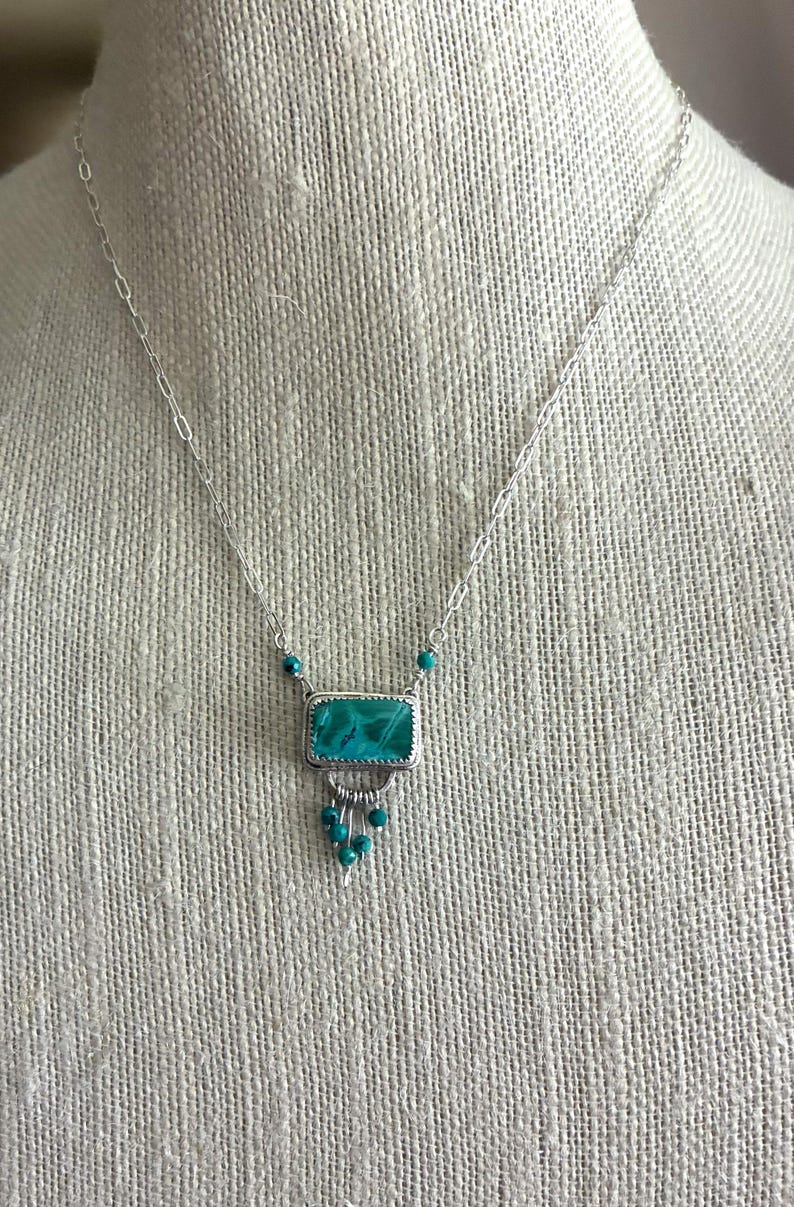May include: A silver necklace featuring a rectangular teal stone pendant set in a serrated bezel. The pendant is accented with small dangling teal beads and is suspended from a delicate silver chain with elongated links and small teal beads.