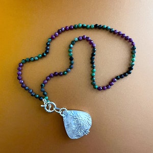May include: A necklace with a silver pendant and a multi-colored beaded chain. The beads are green, purple, and black.