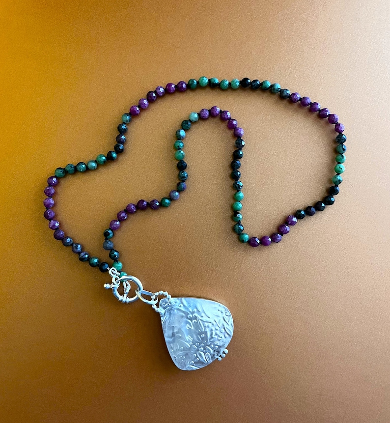 May include: A necklace with a silver pendant and a multi-colored beaded chain. The beads are green, purple, and black.