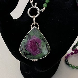 May include: A teardrop-shaped pendant necklace with a green and pink gemstone set in silver. The pendant is attached to a silver chain with a clasp.