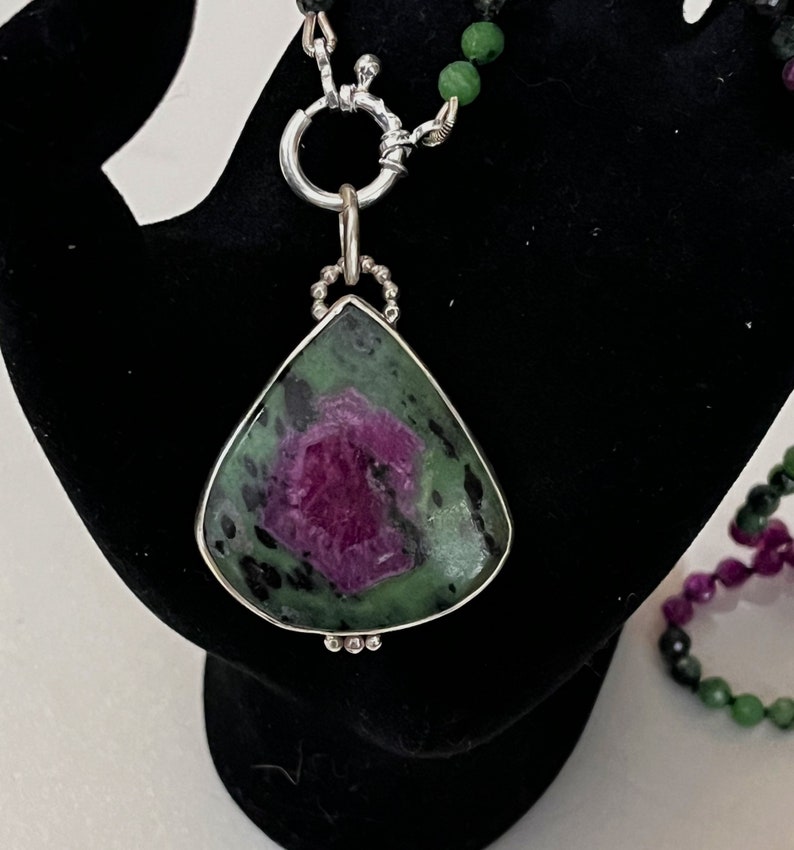 May include: A teardrop-shaped pendant necklace with a green and pink gemstone set in silver. The pendant is attached to a silver chain with a clasp.