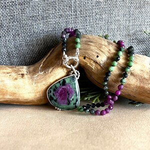 May include: A green, black, and pink gemstone necklace with a large green and pink pendant. The pendant is a teardrop shape and has a silver clasp.