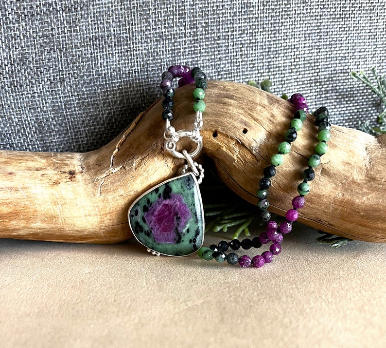 May include: A green, black, and pink gemstone necklace with a large green and pink pendant. The pendant is a teardrop shape and has a silver clasp.