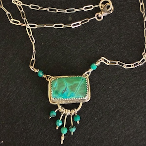 May include: A silver necklace featuring a rectangular green and blue stone pendant set in a serrated metal frame. The pendant is accented with small turquoise beads and a dangling fringe of silver wires tipped with additional turquoise beads.