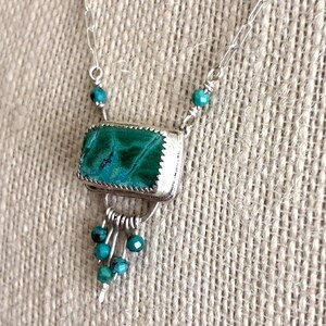 May include: A silver necklace featuring a rectangular malachite pendant with a serrated bezel setting. The pendant is accented with small turquoise beads and a dangling fringe of silver wires tipped with additional turquoise beads.